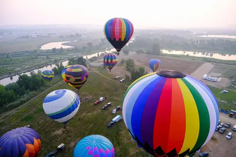 An aerial view shows several colorful hot air balloons flying over a landscape with a river.