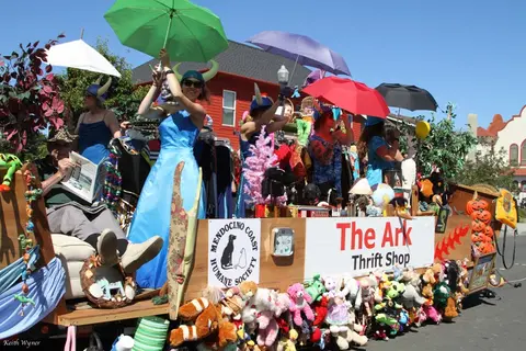 A parade float for a thrift shop features people holding umbrellas and stuffed animals lining the front.
