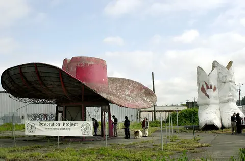 An outdoor view of a giant hat and boots roadside attraction undergoing restoration with people nearby.