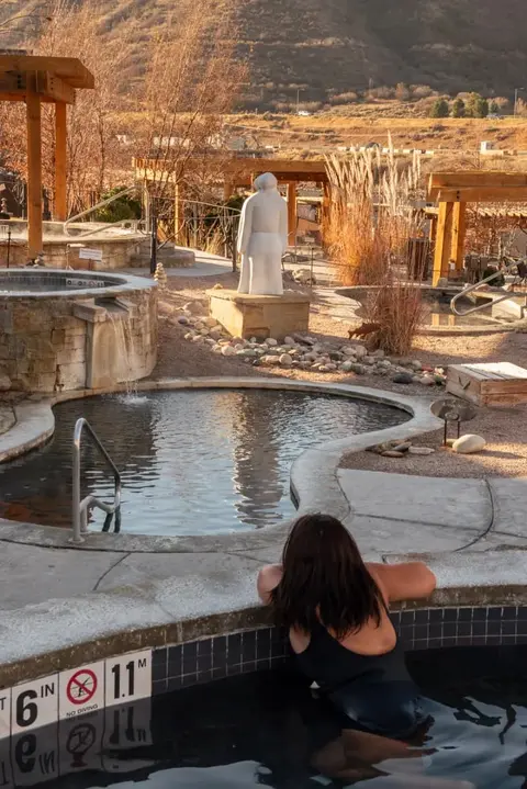 A person relaxing in an outdoor hot spring pool with stone and wood structures around.