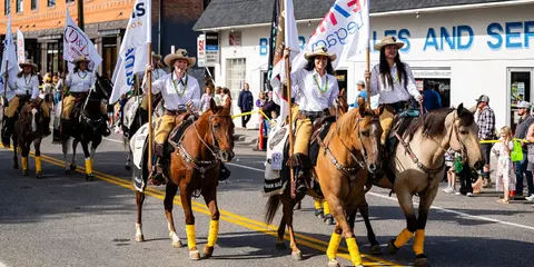 A group of women ride horses carrying flags in a parade down a street.