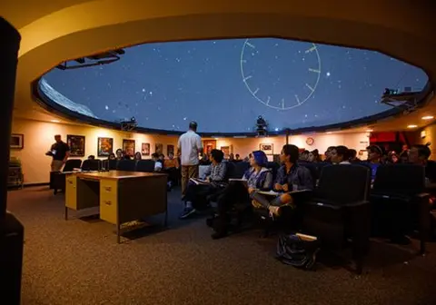 Students seated inside a planetarium watching a presentation on the dome ceiling.