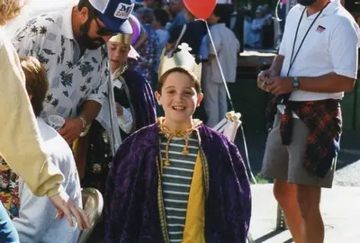 A child wearing a crown and cape stands outdoors among a crowd.