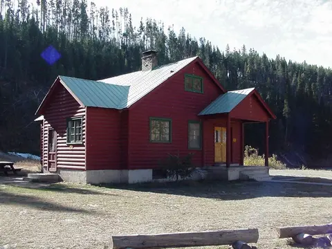 A red log cabin with a green roof stands in a clearing near a forest.