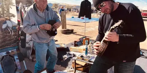 An older man stands near a table of items while another man plays a stringed instrument outdoors.