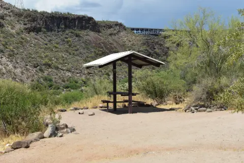 A shaded picnic table sits in a desert landscape with hills and a bridge in the background.