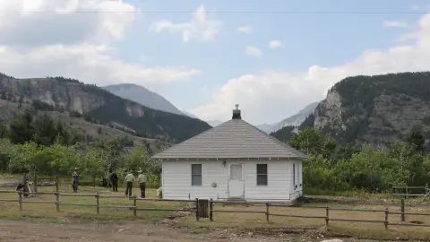A small white building sits in a valley surrounded by mountains and trees.