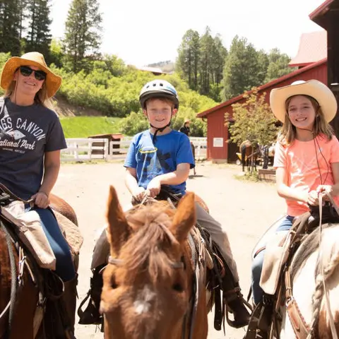 Three people ride horses on a sunny day near a red barn.