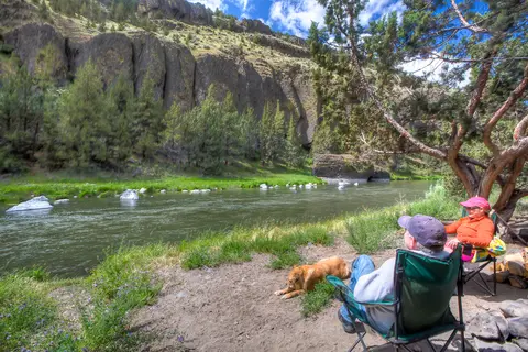 Two people and a dog relaxing in camping chairs by a river near rocky cliffs.