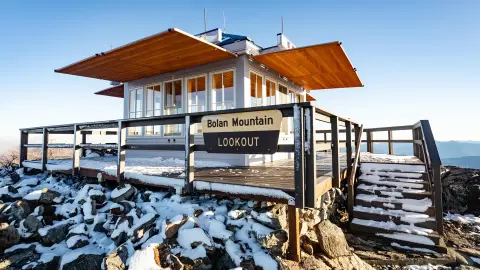A lookout building sits on a snowy mountain peak with a sign on the railing.