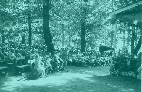 A historic photograph showing a crowd of people seated outdoors under trees.