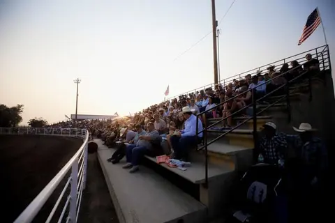 A crowd of people seated in outdoor bleachers watching an event near a dirt arena.