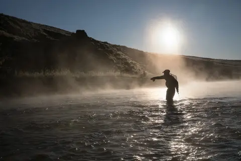 A person fly fishes in a river during a sunny morning with mist rising off the water.