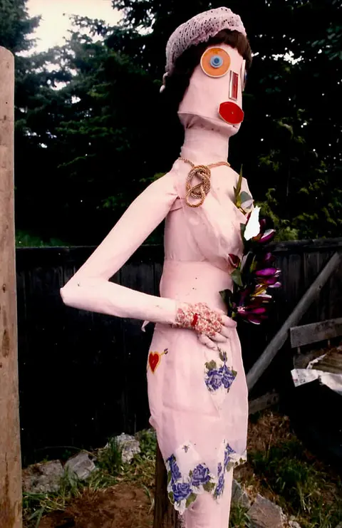 A homemade doll-like mannequin figure stands outdoors holding artificial flowers.