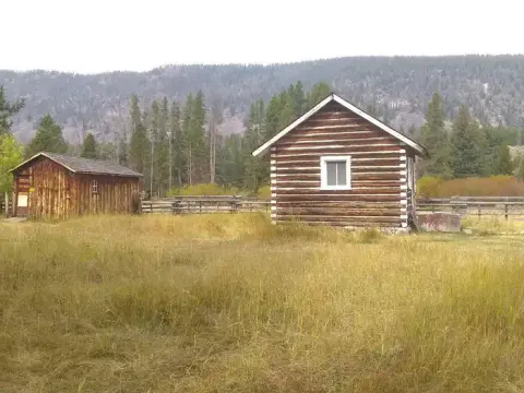 Two rustic wooden cabins sit in a grassy field with trees and mountains in the background.