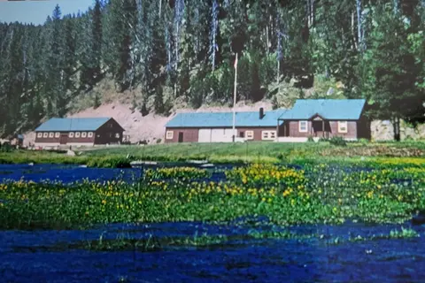 An exterior view of rustic buildings near water with a forested hillside in the background.