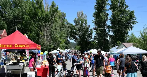 A crowd of people walks along a street lined with vendor tents and trees.