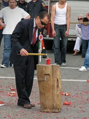 A man in a suit lights a firecracker on a tree stump during an outdoor event.