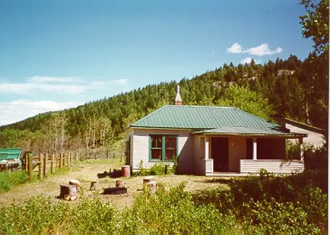 A single-story house with a green roof sits in front of a tree-covered hill.