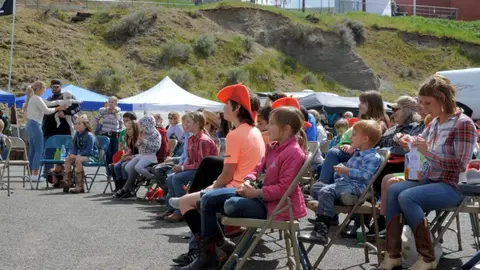 A crowd of children and adults seated outdoors watching an event.