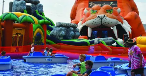 Children ride in small blue paddle boats in front of large inflatable structures.