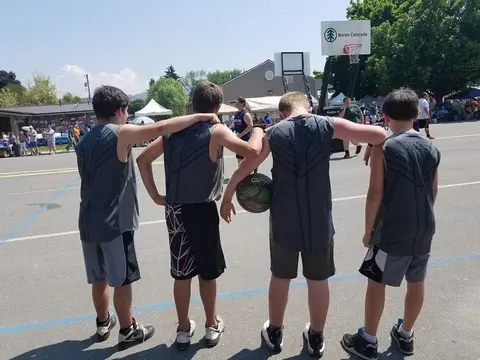 Four young basketball players stand together facing away from the camera on an outdoor court.
