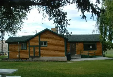 A single-story wooden cabin with a green roof stands in a grassy area.