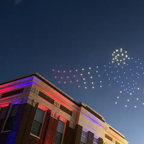 A drone light show flies in the night sky over a lit building.
