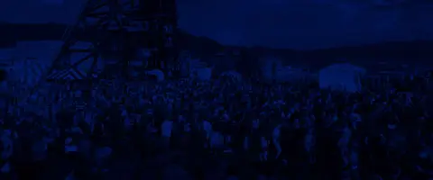 A large crowd of people gathers outdoors near industrial structures under a blue filter.