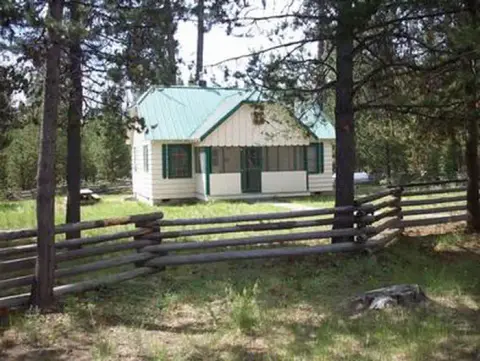A cabin with a green roof stands behind a rustic wooden fence among pine trees.