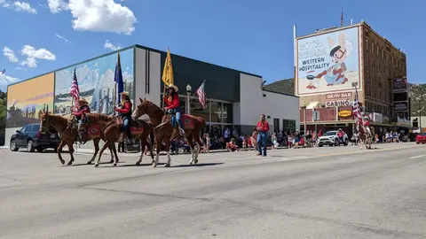 People ride horses carrying flags in a street parade past crowds and buildings.