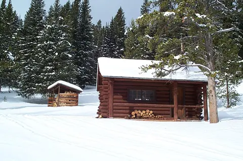 A small log cabin and woodshed in a snowy forest.