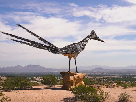A large roadrunner sculpture made of metal stands on a pedestal overlooking a desert valley and mountains.