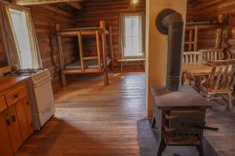 An interior view of a rustic log cabin with wooden bunk beds, a stove, and a dining table.