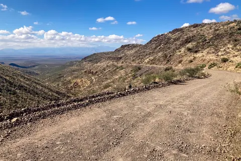 A dirt road winding through a mountainous desert landscape under a blue sky with clouds.