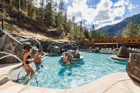 People swim and relax in outdoor pools surrounded by rocks and forested mountains.