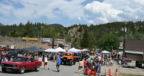 A crowd gathers along a street lined with vendor tents and parked classic cars.