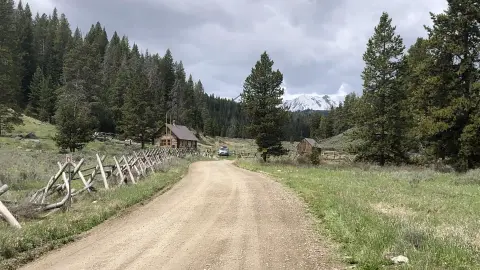 A dirt road leading past a wooden cabin toward snow-capped mountains and a forest.