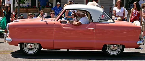 A vintage pink and white car drives down a street past a crowd of people.