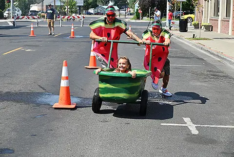 People dressed in watermelon costumes push a child in a decorated tub along a street.