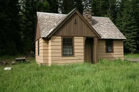 A small wooden cabin stands in a grassy clearing surrounded by trees.