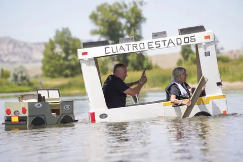 Two people ride in a homemade cardboard boat on a river.