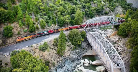 A train crosses a metal bridge over a river surrounded by forested hills.
