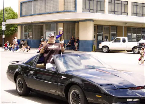 A person waving from a black convertible car in a street parade.