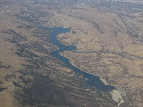An aerial view shows a long, winding reservoir nestled among dry, rolling hills.
