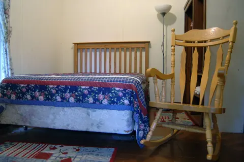 A wooden rocking chair sits in a bedroom with a made bed.