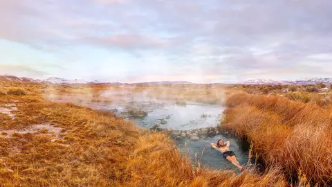 A person relaxing in a natural hot spring surrounded by grassy fields and distant mountains.