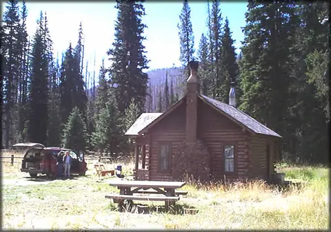 A log cabin stands in a clearing surrounded by trees, with a picnic table and a vehicle nearby.
