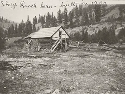 Historic black and white photo of a log building in a mountainous landscape with handwritten text at the top.