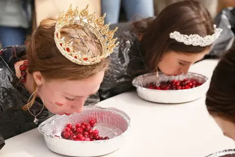 Two people wearing crowns participate in a contest involving bowls of red berries.
